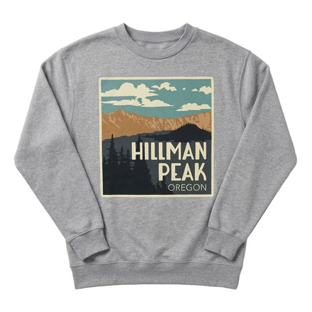 A heather gray crewneck sweatshirt featuring a retro-style graphic print of Hillman Peak, Oregon, with mountains, pine trees, and clouds in muted teal, tan, cream, and dark gray tones.