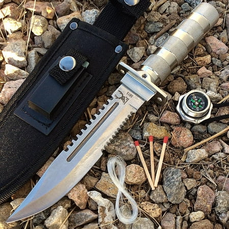 A fixed-blade knife with a silver serrated blade and textured silver handle, shown with a black sheath on a bed of small rocks. A compass, three wooden matches with red tips, and a coiled cord are arranged nearby.