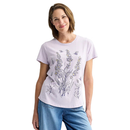 A woman wearing a light lavender crewneck t-shirt with a botanical line-art print of wildflowers and bees, paired with blue jeans, standing against a plain light gray wall. 