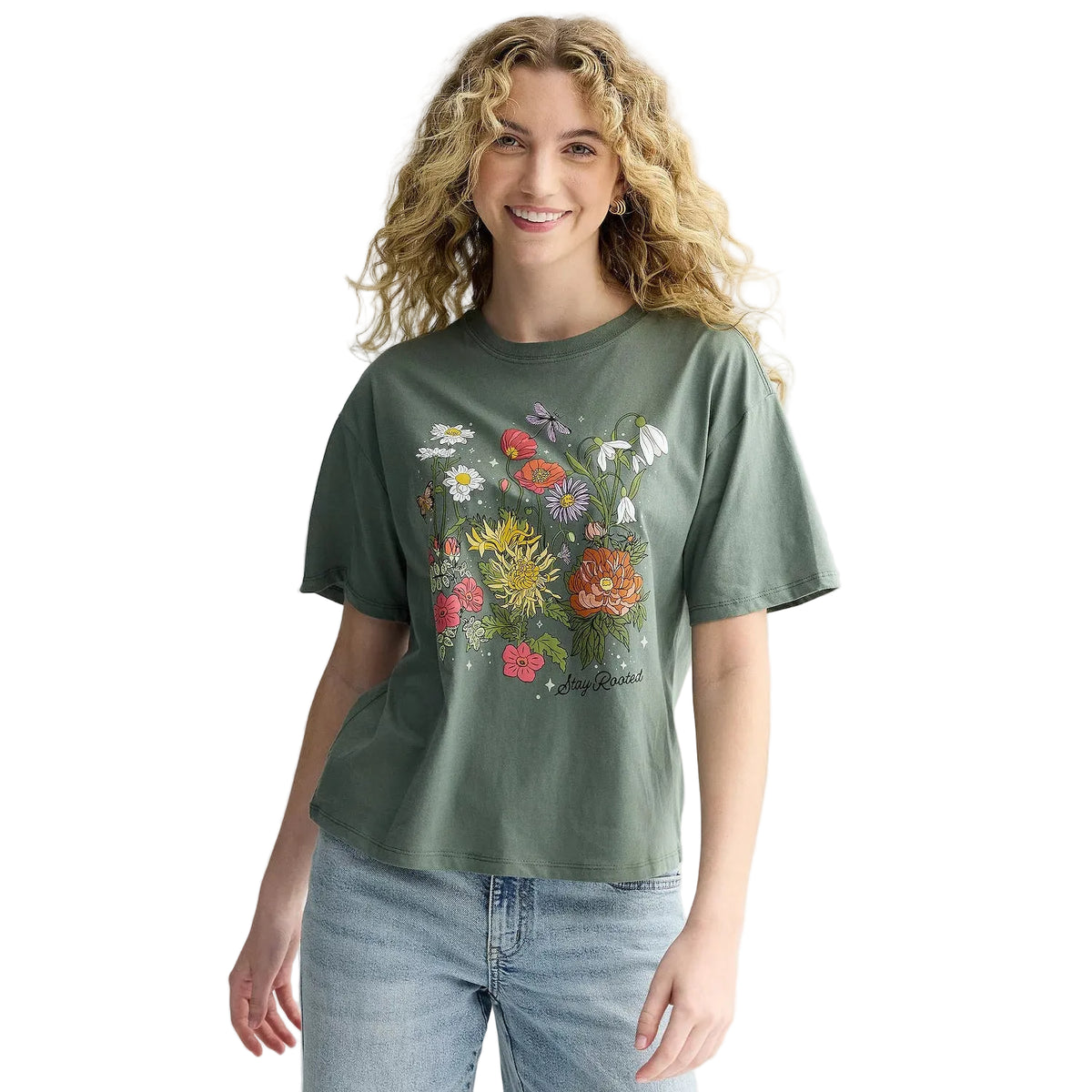 A woman with curly blonde hair wearing a sage green short-sleeve t-shirt with a colorful wildflower graphic on the front, paired with light blue jeans, against a plain light gray background.