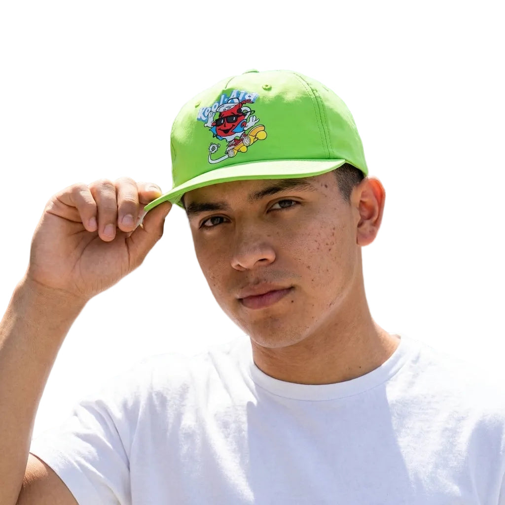 A man wearing a bright lime green baseball cap with a colorful cartoon monster graphic on the front, holding the brim with his hand. He is wearing a white t-shirt and posing outdoors against a blurred modern building background.