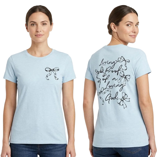 A woman modeling a light blue short-sleeve t-shirt, showing both the front and back views. The front features a small black bow graphic on the left chest, and the back features a larger black graphic with the words 'Living Proof of a Loving God' surrounded by bow illustrations.