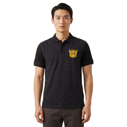 A man wearing a short-sleeve polo shirt with a black and dark gray grid pattern, solid black sleeves, and a yellow Decepticon insignia on the left chest, standing against a plain off-white background.