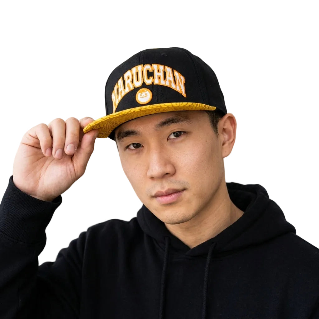 A man wearing a black snapback cap with gold trim and the word 'MARUCHAN' in gold letters, posing in a black hoodie in front of a modern glass building.
