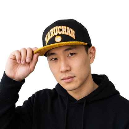 A man wearing a black snapback cap with gold trim and the word 'MARUCHAN' in gold letters, posing in a black hoodie in front of a modern glass building.