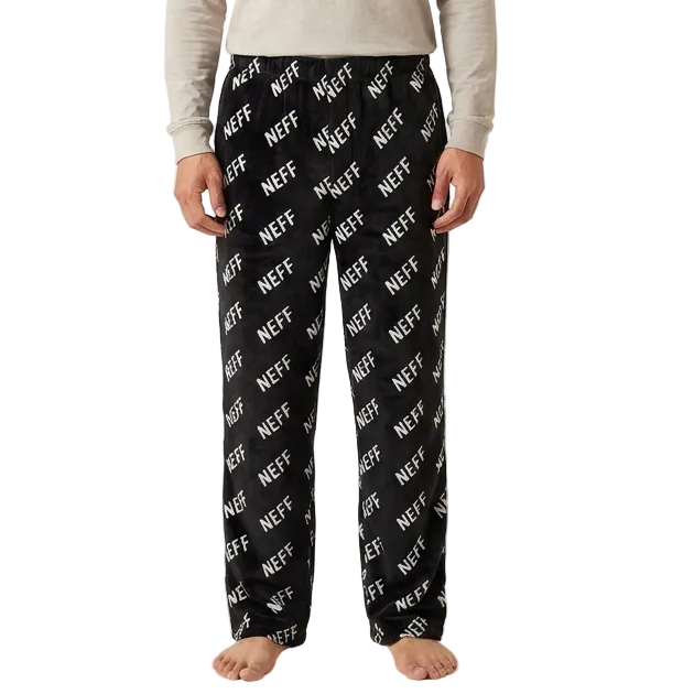 A man stands against a plain light beige studio background wearing a light beige long-sleeve crewneck shirt and black pajama pants with a white diagonal 'NEFF' logo print.