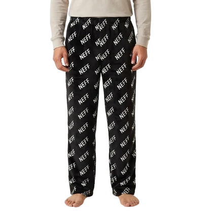 A man stands against a plain light beige studio background wearing a light beige long-sleeve crewneck shirt and black pajama pants with a white diagonal 'NEFF' logo print.
