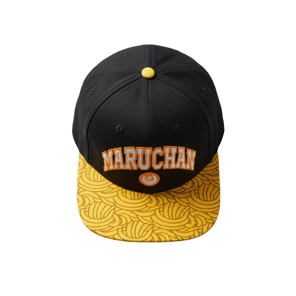 A black flat-brim cap with a yellow patterned visor and yellow button, featuring the word 'MARUCHAN' in orange arched letters with a small orange circular logo. The cap is styled on a light gray surface with chopsticks and a white bowl in the background.