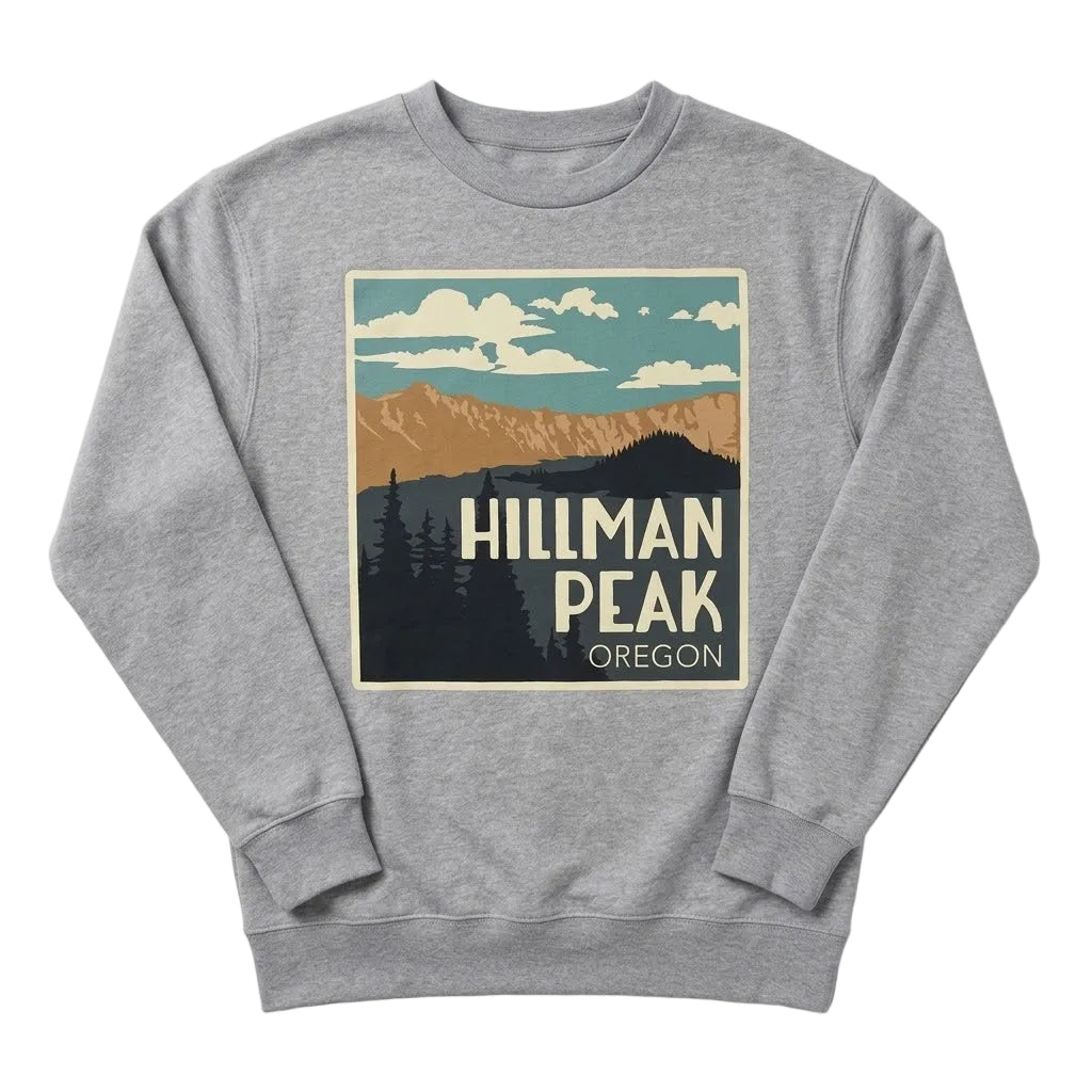 A heather gray crewneck sweatshirt featuring a retro-style graphic print of Hillman Peak, Oregon, with mountains, pine trees, and clouds in muted teal, tan, cream, and dark gray tones. 